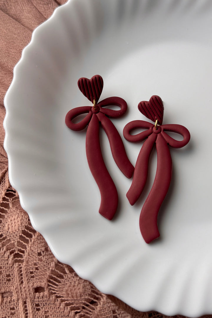 Red bow-shaped earrings on a white plate with a brown textured surface underneath.