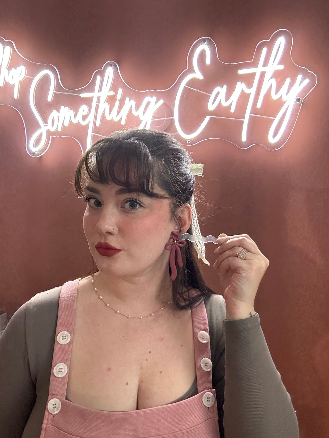 Woman in pink overalls holding a cigarette with 'Something Earthy' neon sign in the background