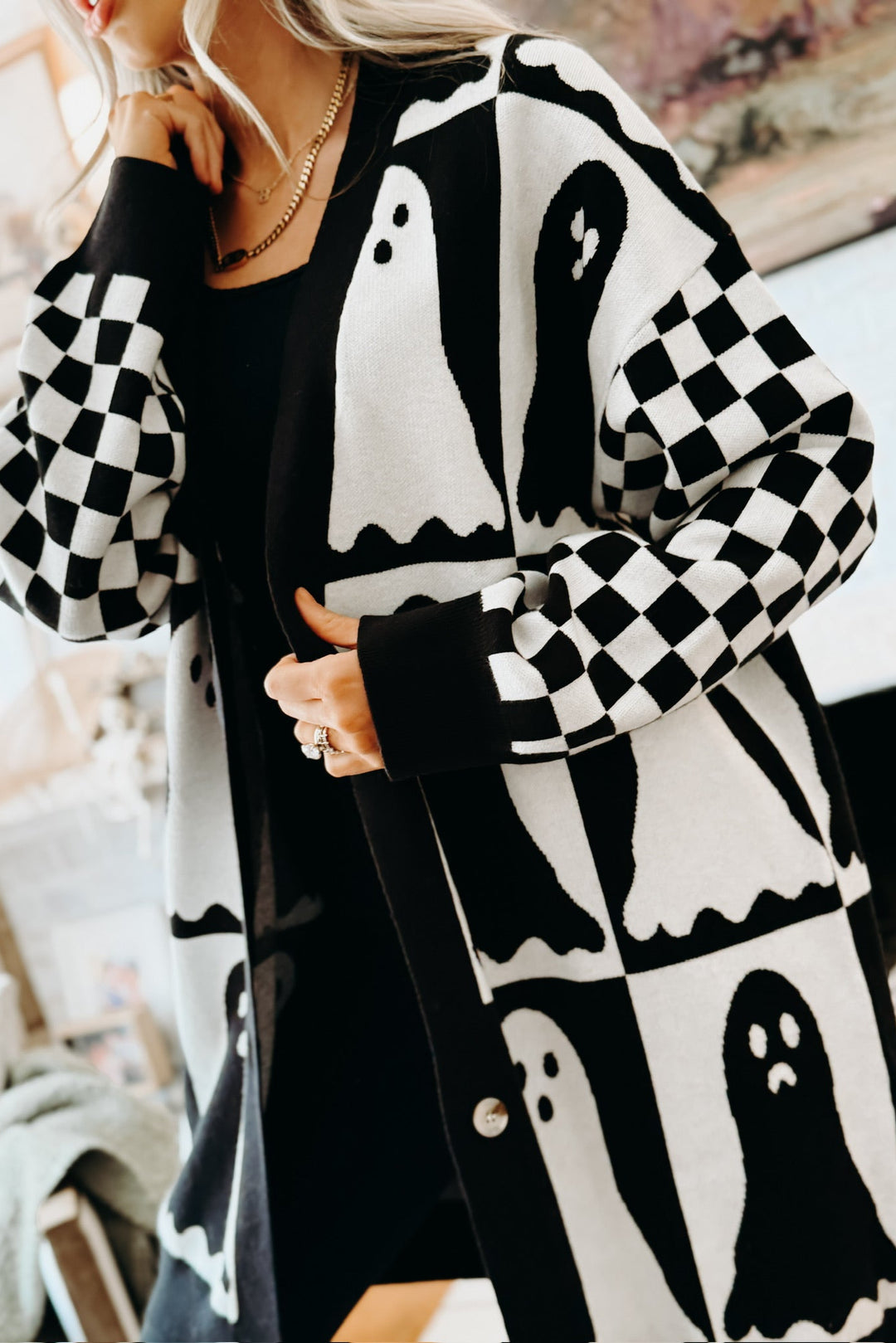 A woman wearing a black and white cardigan with ghost and checkerboard patterns over a black dress, in a softly lit indoor setting.