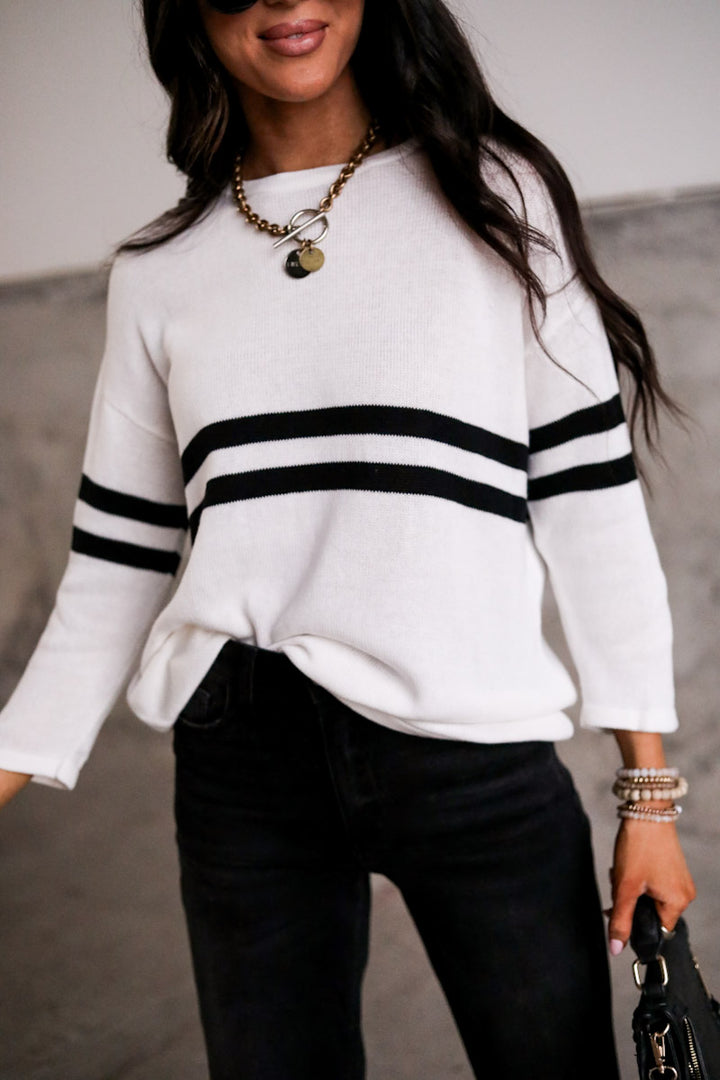 Person wearing a white sweater with black stripes and black pants, holding a handbag.