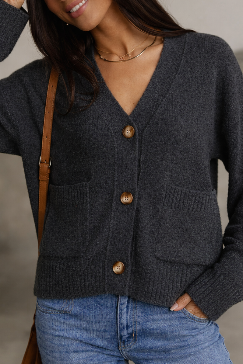 Woman wearing a dark gray cardigan with brown buttons and blue jeans, standing against a neutral background.