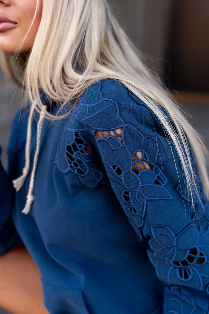 Close-up of a woman wearing a blue hoodie with floral eyelet lace detailing on the sleeves and white drawstrings.