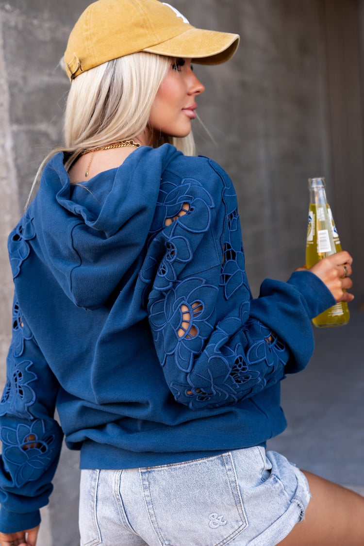A woman wearing a blue floral embroidered hoodie with cutout details, a yellow baseball cap, and denim shorts, holding a glass of beer against a gray concrete wall.