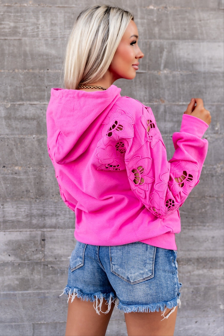 A woman wearing a bright pink cropped hoodie with butterfly cutout details on the sleeves, paired with denim shorts, standing against a gray concrete wall.
