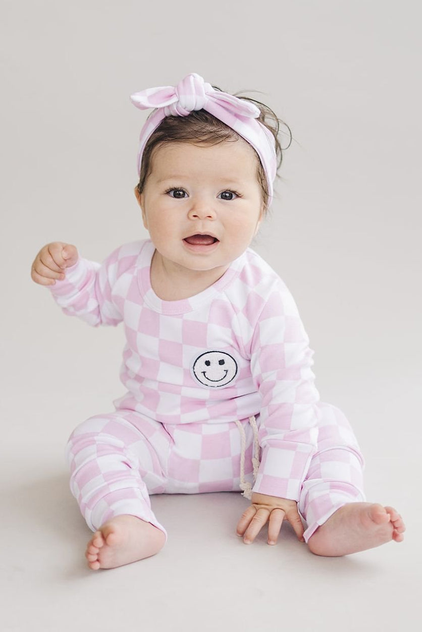 Baby wearing a pink checkered outfit with a smiley face design on a plain background